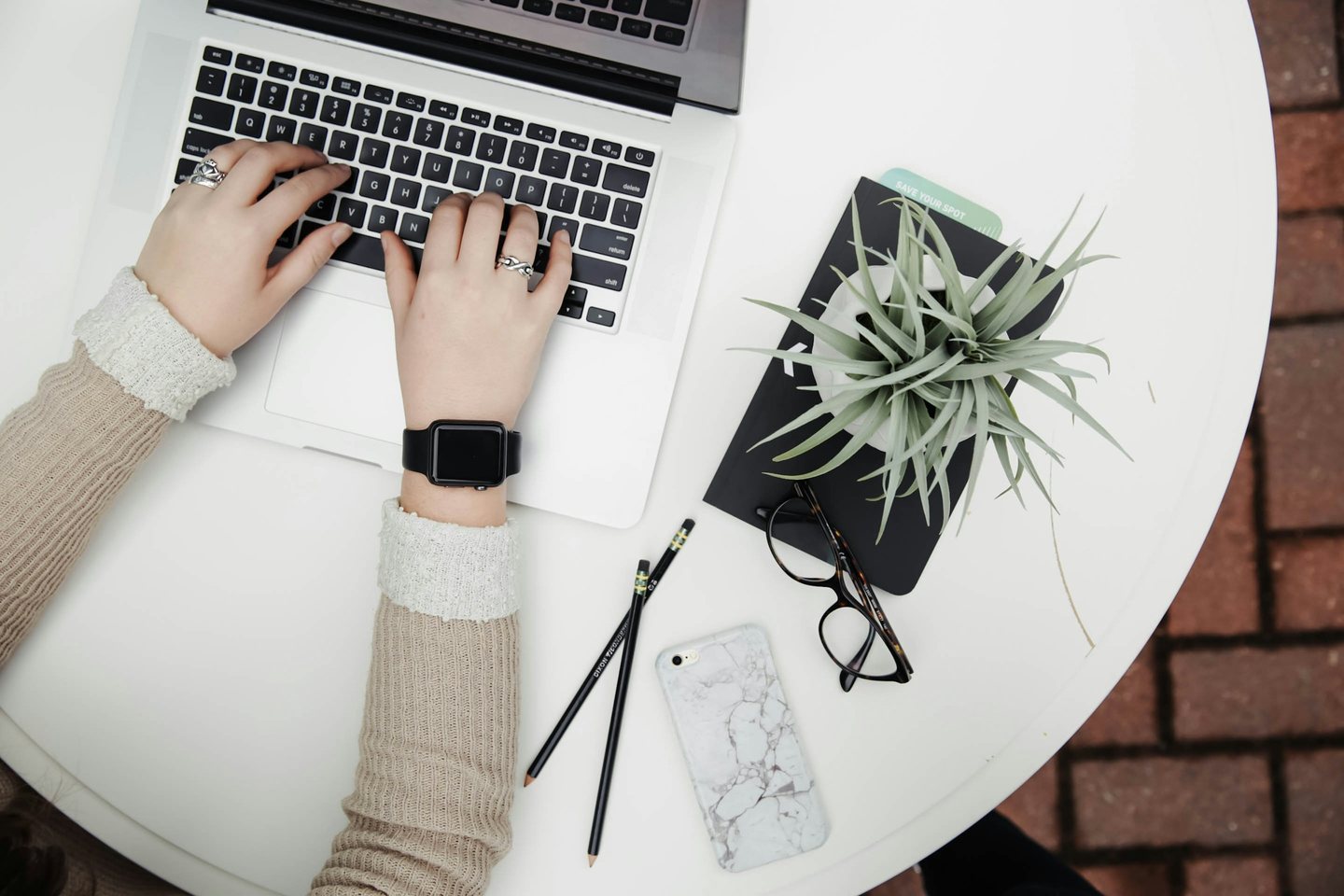 Hands typing on a laptop at a round white desk beside a plant and glasses