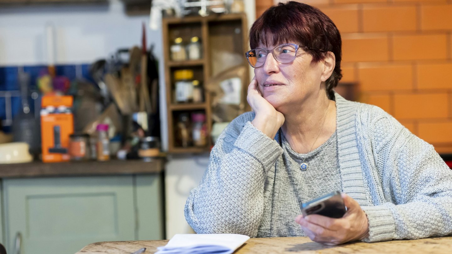 An older woman at her kitchen table holding her phone, thinking
