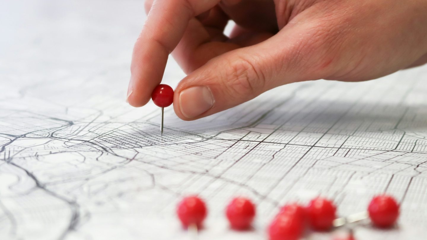 A hand pressing a red pin into a city street map, with more red pins beside it