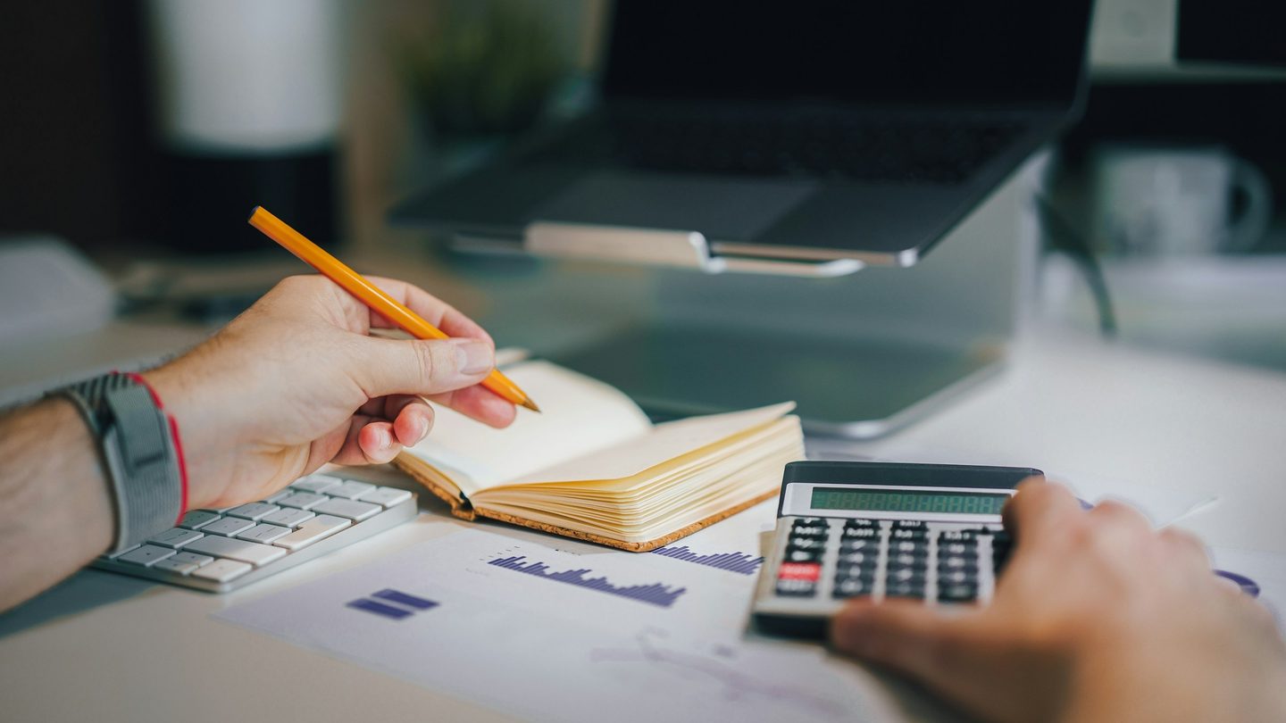 A person writing in a notebook beside a calculator and a printed chart